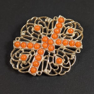 May include: A gold-toned brooch with an openwork filigree design. The brooch features a cross shape adorned with orange, round beads. The cross is centered on the filigree, creating a decorative and ornate accessory.