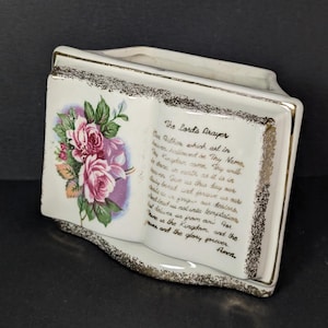 May include: A cream-coloured ceramic planter shaped like an open book with gold-trimmed edges. The left page features a pink rose illustration, while the right page displays The Lord's Prayer in gold script. The piece has a hollow centre for storage.