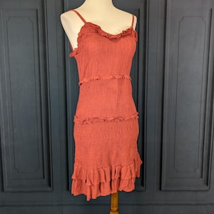 Puede incluir: Un vestido color terracota con tirantes finos y volantes. El vestido presenta una tela texturizada y varias filas de volantes. Se exhibe en un maniquí de madera, mostrando su diseño y ajuste.