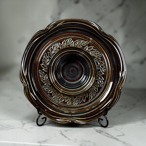May include: A handcrafted ceramic plate with a scalloped edge. The plate features a dark brown and burgundy glaze with a white leaf design around the center. The plate is displayed on a black metal stand.