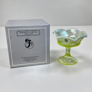 May include: A yellow-green Fenton Art Glass Company compote with iridescent highlights. The compote has a ruffled rim and sits on a pedestal base. A gray box with the text "HANDMADE IN AMERICA SINCE 1905" is next to the compote.