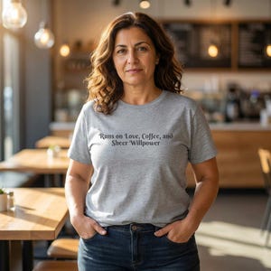 Puede incluir: Una camiseta gris claro con el texto "Runs on Love, Coffee, and Sheer Willpower" en escritura gris oscuro. La camiseta se combina con vaqueros azules. La persona está en un café.