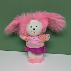 May include: A plush toy with a pink furry head and ears, a white face, and a small black nose and eyes. It wears a pink shirt with the text "I Love Cheerleading" and a purple skirt. The toy has pink feet and arms.