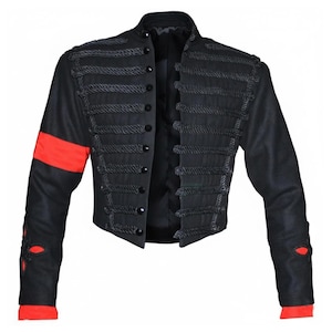 Puede incluir: Chaqueta corta negra con cuello mandarín, adornada con detalles de cuerda oscura y botones negros. Presenta una franja roja en la manga izquierda y detalles rojos en los puños. Prenda de inspiración militar.