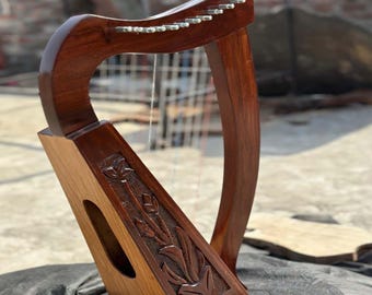 Puede incluir: Un arpa de madera hecha a mano con un acabado marrón oscuro. El arpa presenta un diseño floral tallado en la caja de resonancia y clavijas de afinación metálicas. Las cuerdas son visibles, sobre un fondo exterior borroso.