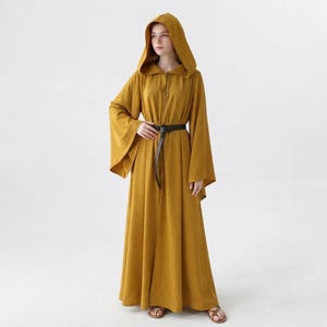 Pode incluir: Uma túnica longa até ao chão de cor amarelo mostarda com capuz, mangas compridas e largas e fecho de correr frontal. A peça é ajustada na cintura com um cinto preto fino e combinada com sandálias castanhas simples.