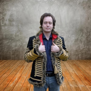 Op de afbeelding: Een zwarte jas in militaire stijl met gouden biezen en een kraag van imitatiebont. De jas heeft een rode kraag en gouden kwastjes. De persoon draagt een blauw overhemd en een blauwe jeans.