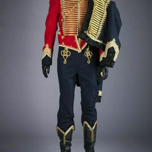 Könnte beinhalten: Eine rot-blaue Militäruniform mit goldenen Verzierungen und einem schwarzen Pelzkragen. Die Uniform besteht aus einer Jacke, einer Hose und Stiefeln.