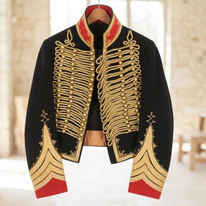 Puede incluir: Una chaqueta de estilo militar negra con detalles de cordón trenzado dorado en la parte delantera, un cuello alto rojo con bordados dorados y puños rojos con detalles de galones dorados. La chaqueta está abierta y cuelga de una percha.