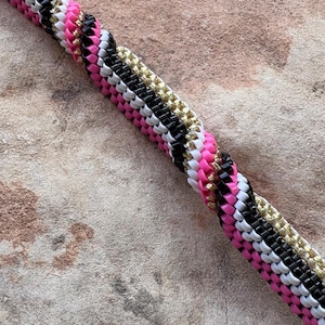 May include: Close-up of a braided cord with alternating sections of pink, white, black, and gold. The cord is made of small, tightly woven beads, creating a textured surface. The background is a textured, neutral-toned surface.