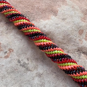May include: A colorful, braided bracelet featuring a repeating pattern of red, black, neon green, and coral-colored segments. The bracelet is tightly woven, creating a textured, spiral design. The background is a neutral, textured surface.