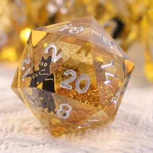 以下が含まれることがあります： 金色でキラキラした内装の多面体サイコロのセット。 d20サイコロに焦点を当て、黒猫のイラストとシルバーの数字を表示しています。 サイコロはテーブルゲーム用に設計されています。
