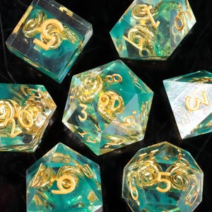 May include: A set of seven polyhedral dice. Each die is a clear teal color with gold-colored numbers and a gold swirl design inside. The dice are arranged on a dark surface.