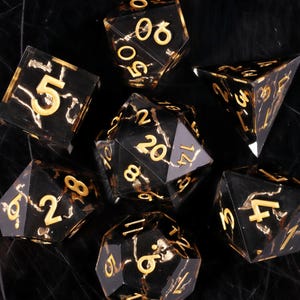 Puede incluir: Juego de siete dados poliédricos con un diseño veteado negro y dorado. Cada dado presenta números dorados. Los dados están diseñados para juegos de rol de mesa y otros juegos de dados.