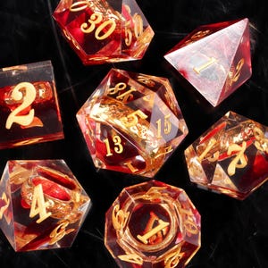 May include: Set of seven polyhedral dice with a red and gold color scheme. The dice are transparent with gold-colored numbers and decorative elements inside. The dice are arranged on a black surface.