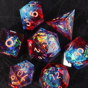 May include: A set of seven polyhedral dice with a vibrant, colorful design. The dice feature a mix of red and blue hues with iridescent accents, and each die has gold-colored numbers. The dice are designed for tabletop gaming.