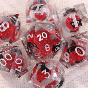 Puede incluir: Un conjunto de dados transparentes de múltiples caras con interiores de purpurina roja y numeración blanca. Algunos dados presentan siluetas de gatos negros. Los dados están diseñados para juegos de mesa y juegos de rol.