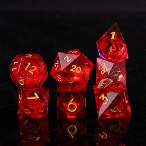 May include: A set of seven red, transparent polyhedral dice with gold numbering. The dice are arranged on a black surface, showcasing their various geometric shapes, including a cube, a d20, and a d4. The dice are designed for tabletop games.