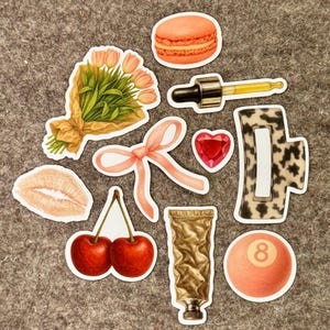 Könnte beinhalten: Eine Sammlung von Aufklebern mit verschiedenen Designs. Enthält einen Tulpenstrauß, eine rosa Schleife, einen roten herzförmigen Edelstein, ein Kirschenpaar, eine Tube Farbe, ein Macaron, eine Pipette, einen Lippenabdruck, eine Haarspange und eine Billardkugel.