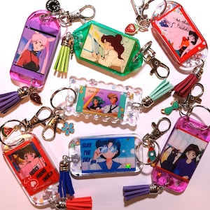 May include: A set of nine colourful keychains featuring anime characters and inspirational phrases. Each keychain has a clear plastic charm with a different image and text, such as "Ride or Die", "Be That Baddie", and "Slay the Day". The keychains also have colourful tassels and charms.