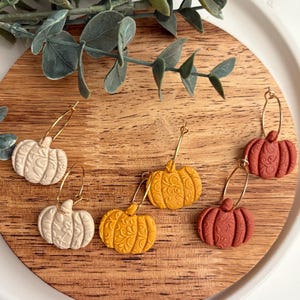 May include: A collection of pumpkin-shaped earrings in cream, yellow, and rust-red colors. Each earring features a detailed floral pattern and is suspended from a gold-colored hoop. The earrings are displayed on a wooden cutting board.