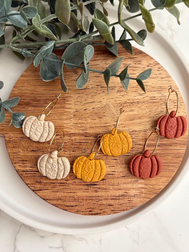 May include: A collection of pumpkin-shaped earrings in cream, yellow, and rust-red colors. Each earring features a detailed floral pattern and is suspended from a gold-colored hoop. The earrings are displayed on a wooden cutting board.