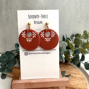 May include: Terracotta-colored, circular earrings with white floral cutouts. The earrings are displayed on a white card with the text "SEVENTY-THREE DESIGNS" and a website address. The earrings hang from gold-colored posts.