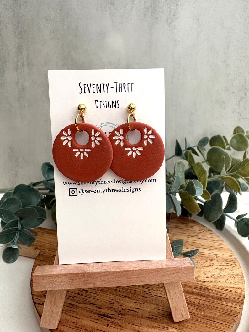 May include: Terracotta-colored, circular earrings with white floral cutouts. The earrings are displayed on a white card with the text "SEVENTY-THREE DESIGNS" and a website address. The earrings hang from gold-colored posts.