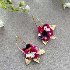 May include: A pair of hoop earrings featuring floral charms with magenta, white, and black petals. Each earring includes two gold-toned metallic leaves with a textured, veined surface attached to the base of the flower, suspended from a thin gold hoop.