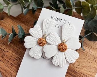 May include: White daisy earrings with textured petals and a gold-colored center. The earrings are displayed on a white card with the text "SEVENTY-THREE DESIGN" and a wooden surface.