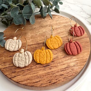 May include: A collection of pumpkin-shaped earrings in cream, yellow, and terracotta colors. The earrings have gold-colored hoops and are displayed on a wooden surface. The pumpkins have detailed embossed patterns.
