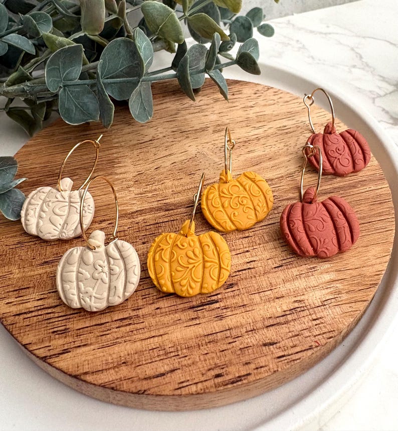 May include: A collection of pumpkin-shaped earrings in cream, yellow, and terracotta colors. The earrings have gold-colored hoops and are displayed on a wooden surface. The pumpkins have detailed embossed patterns.