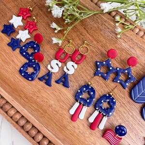 May include: A selection of red, white, and blue patriotic earrings. The designs include star shapes, the letters "USA", and flag-themed elements. The earrings are presented on a wooden tray, with floral decorations.