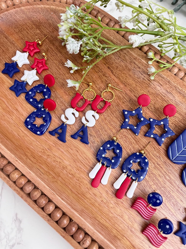 May include: A selection of red, white, and blue patriotic earrings. The designs include star shapes, the letters "USA", and flag-themed elements. The earrings are presented on a wooden tray, with floral decorations.