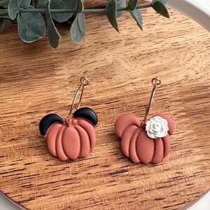 Puede incluir: Un par de pendientes de calabaza de color terracota con aros dorados. Un pendiente tiene acentos de oreja negros, el otro tiene un acento de flor blanca. Los pendientes est&aacute;n sobre una superficie de madera.