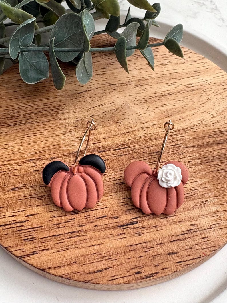 Puede incluir: Un par de pendientes de calabaza de color terracota con aros dorados. Un pendiente tiene acentos de oreja negros, el otro tiene un acento de flor blanca. Los pendientes est&aacute;n sobre una superficie de madera.
