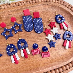 May include: A selection of red, white, and blue earrings with patriotic themes. The earrings feature star and flag designs, some with a rectangular top and chevron pattern. The earrings are displayed on a wooden tray.