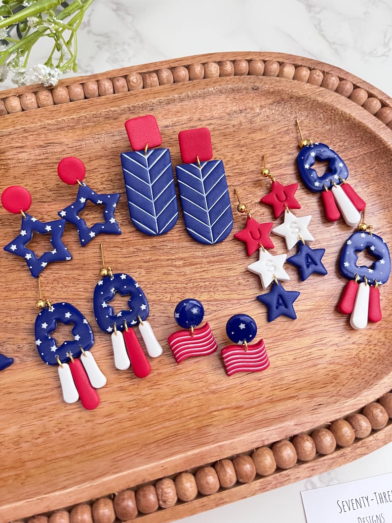 May include: A selection of red, white, and blue earrings with patriotic themes. The earrings feature star and flag designs, some with a rectangular top and chevron pattern. The earrings are displayed on a wooden tray.