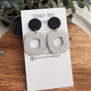 May include: Earrings with a speckled, square design and a black circular top. The earrings are attached to a white card with the text "SEVENTY-THREE DESIGNS" and social media handles. The earrings are displayed on a wooden surface.