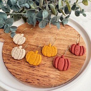 May include: A collection of pumpkin-shaped earrings in cream, yellow, and rust-red hues, displayed on a wooden cutting board. Each earring features a gold hoop and intricate carved details, evoking a fall harvest theme.