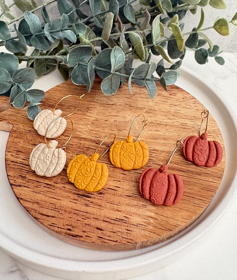 May include: A collection of pumpkin-shaped earrings in cream, yellow, and rust-red hues, displayed on a wooden cutting board. Each earring features a gold hoop and intricate carved details, evoking a fall harvest theme.