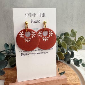 May include: Terracotta-colored, circular earrings with white floral cutouts and gold-colored studs. The earrings are displayed on a white card with the text "SEVENTY-THREE DESIGNS" and social media information. The earrings are on a small wooden easel.