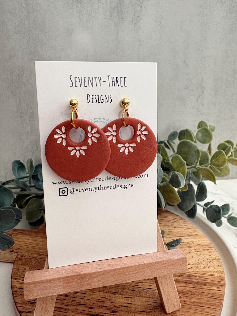 May include: Terracotta-colored, circular earrings with white floral cutouts and gold-colored studs. The earrings are displayed on a white card with the text "SEVENTY-THREE DESIGNS" and social media information. The earrings are on a small wooden easel.