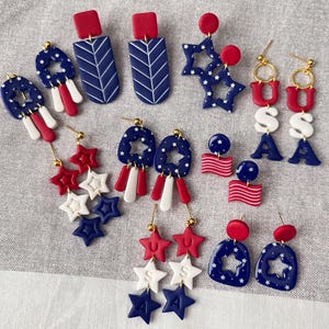 May include: A selection of red, white, and blue earrings with patriotic themes. Designs include star shapes, rectangular shapes with chevron patterns, and earrings spelling out "USA". The earrings are made of polymer clay.