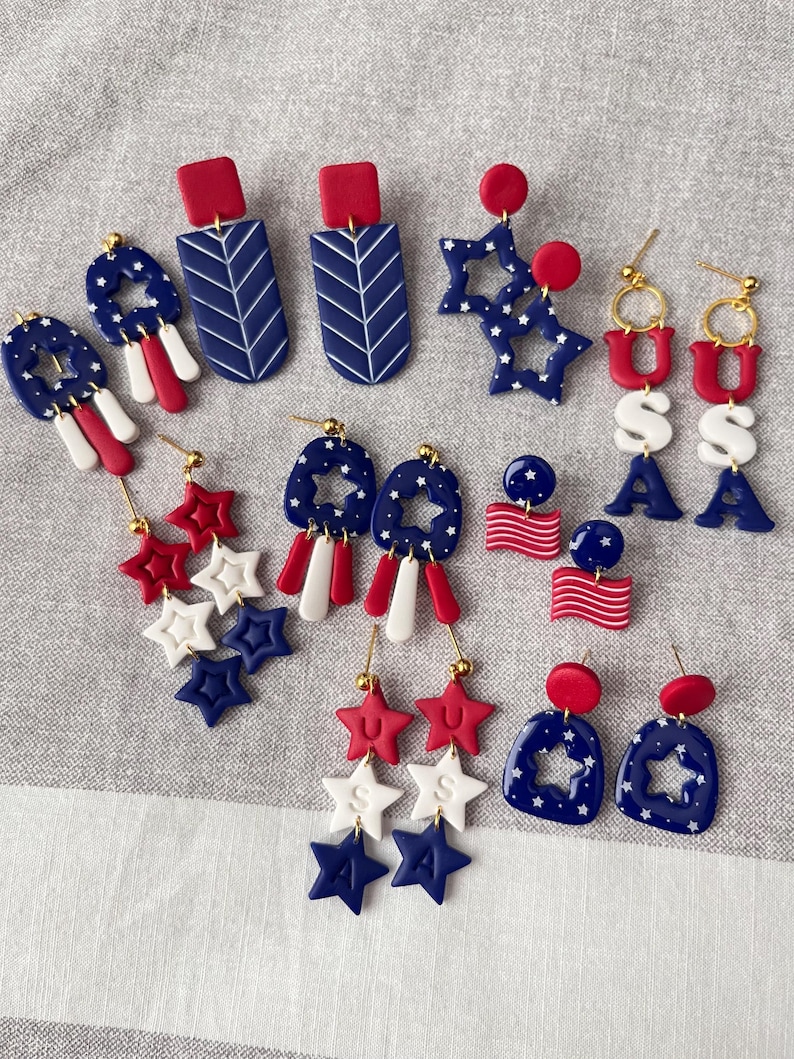 May include: A selection of red, white, and blue earrings with patriotic themes. Designs include star shapes, rectangular shapes with chevron patterns, and earrings spelling out "USA". The earrings are made of polymer clay.
