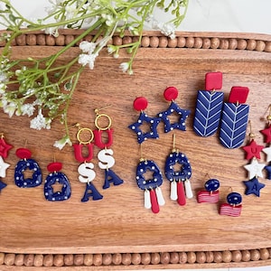 May include: A selection of red, white, and blue earrings with patriotic themes, displayed on a wooden tray. The earrings feature star, USA, and abstract designs. Some have gold-coloured hooks. A sprig of white flowers is in the upper left corner.
