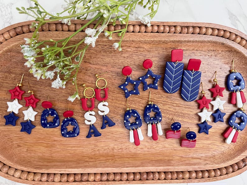 May include: A selection of red, white, and blue earrings with patriotic themes, displayed on a wooden tray. The earrings feature star, USA, and abstract designs. Some have gold-coloured hooks. A sprig of white flowers is in the upper left corner.