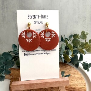 May include: Terracotta-colored, circular earrings with white floral designs and gold-colored posts. The earrings are displayed on a white card with the text "SEVENTY-THREE DESIGNS" and a website address. The earrings are on a small wooden easel.