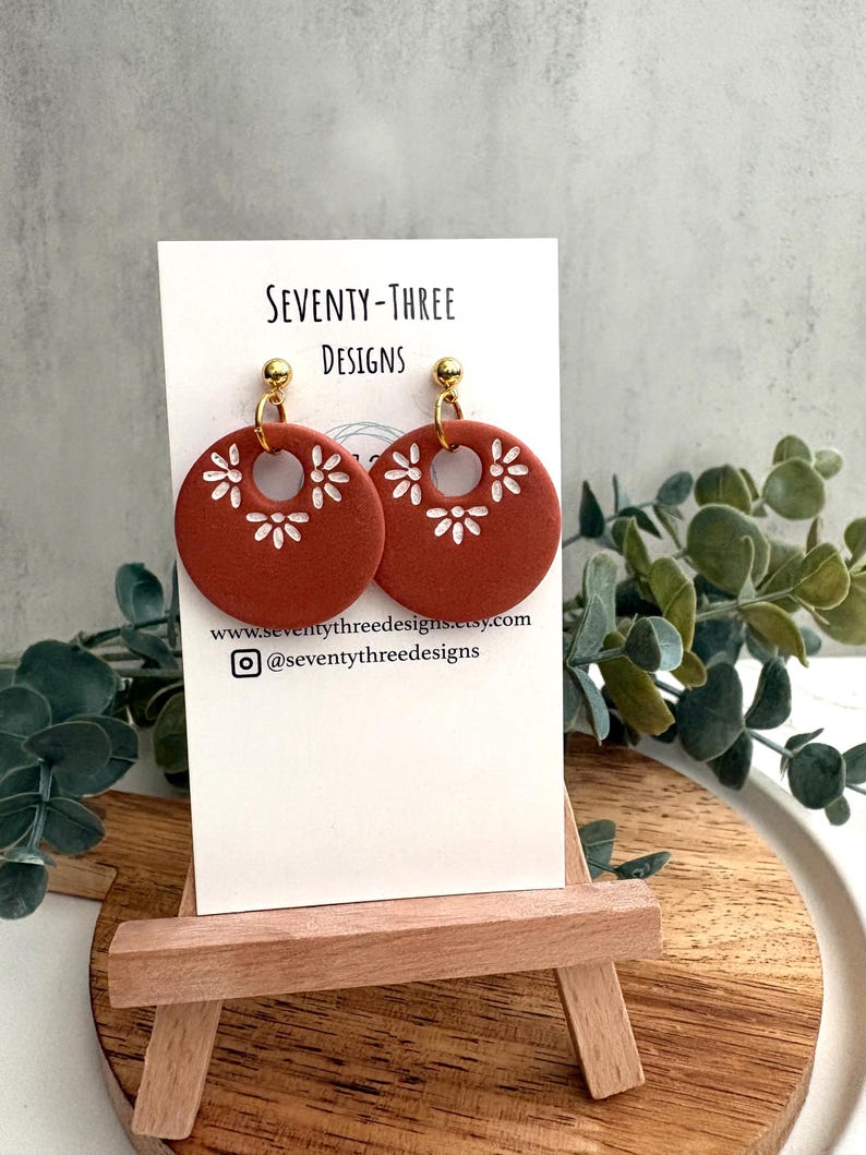 May include: Terracotta-colored, circular earrings with white floral designs and gold-colored posts. The earrings are displayed on a white card with the text "SEVENTY-THREE DESIGNS" and a website address. The earrings are on a small wooden easel.