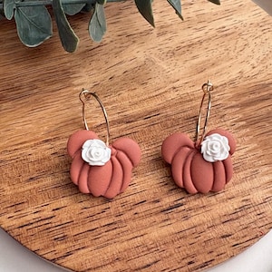 Puede incluir: Un par de pendientes con forma de calabaza de color terracota con detalles de rosas blancas. Los pendientes est&aacute;n sujetos a cierres de aro dorados. Los pendientes se exhiben sobre una superficie de madera.